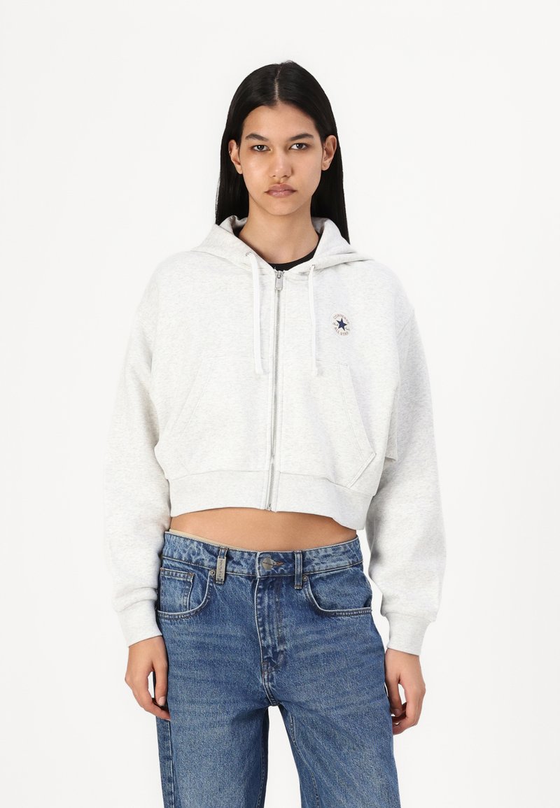 Grey cropped hoodie with a zipper and drawstrings, featuring a small logo on the chest and two front pockets. Worn with blue denim jeans.