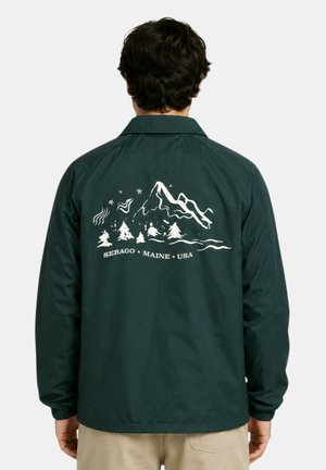 Uomo che indossa una giacca verde scuro con grafica bianca di montagna e albero e testo "SEBAGO MAINE USA" sulla schiena, in piedi davanti a uno sfondo uniforme.
