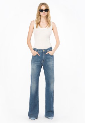 Donna che indossa occhiali da sole neri a forma di occhio di gatto, top senza maniche bianco a costine, jeans blu a gamba larga con le mani nelle tasche, in piedi su uno sfondo bianco.