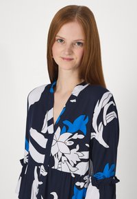 Vestido floral em azul-marinho, com grandes estampas botânicas brancas e azuis, decote em V e mangas com babados. Tecido suave e leve.