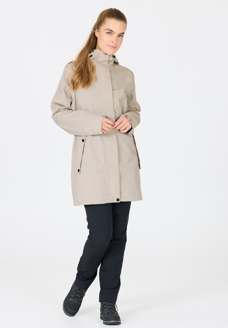 Veste imperméable beige avec capuche, poignets ajustables et deux poches avant. Portée sur un pantalon noir et des bottes de randonnée grises. Texture lisse.