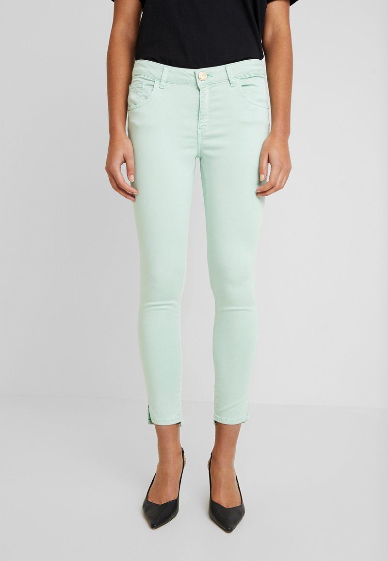 Mint green skinny jeans made from stretch denim, featuring front pockets, a button closure, and subtle hem slits. Worn with black heels.