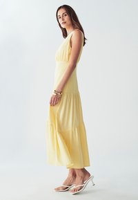 Rumena, brezrokavna maksi obleka s prilegajočim telesom, večplastnim volančkom in lahkotnim materialom. Bele sandale z obelkom dopolnjujejo videz.