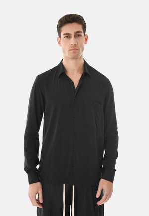 Giovane in piedi immobile che indossa una camicia nera con bottoni e tasca e pantaloni neri con coulisse bianche su sfondo bianco.