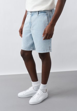 Férfi világoskék farmer rövidnadrágban, fehér zokniban és fehér tornacipőben, semleges háttér előtt áll.