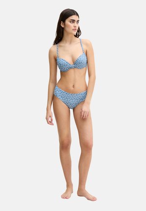 Woman standing barefoot wearing a blue bikini with white floral pattern, looking to the side against a plain background.