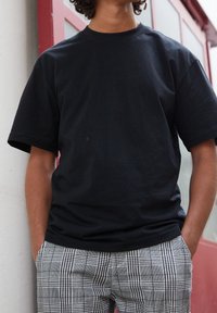 Osoba w gładkiej, czarnej koszulce z krótkim rękawem i czarno-białych, w kratkę spodniach stoi z rękami w kieszeniach.