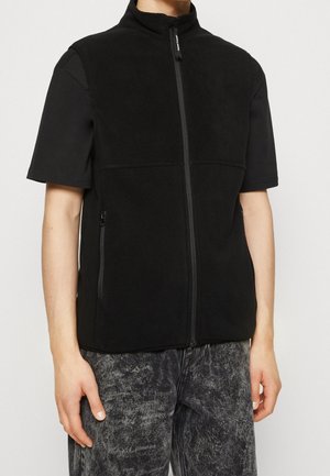 Personne portant un gilet zippé sans manches en polaire noire, sur un t-shirt noir à manches courtes et un jean gris foncé délavé à l'acide.