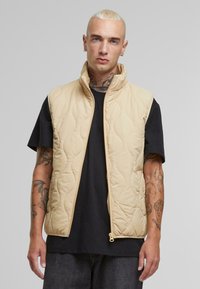 Beige quilted vest with high collar, front zipper, and side pockets, worn over a black shirt, featuring a smooth texture and minimalistic design.