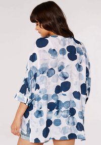 Blouse légère et ample de couleur blanche avec des motifs circulaires bleus variés. Équipée de manches trois-quarts et de fentes latérales pour faciliter les mouvements.