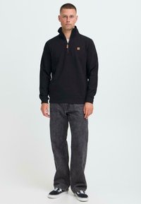 Felpa nera con zip e collo alto, patch logo sul petto. Abbinata a jeans grigi dal taglio morbido e sneakers nere. Design minimalista.