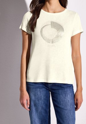 Kobieta w kremowym t-shircie z krótkim rękawem z okrągłym wzorem promieni słonecznych i napisem "SOLEIL", w połączeniu z niebieskimi dżinsami.