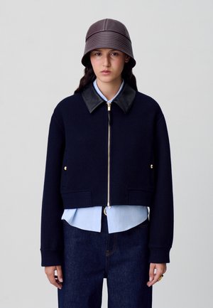 Veste cropped navy avec col en cuir, accessoires dorés et fermeture éclair, superposée à une chemise bleu clair et un jean en denim foncé. Chapeau seau marron.