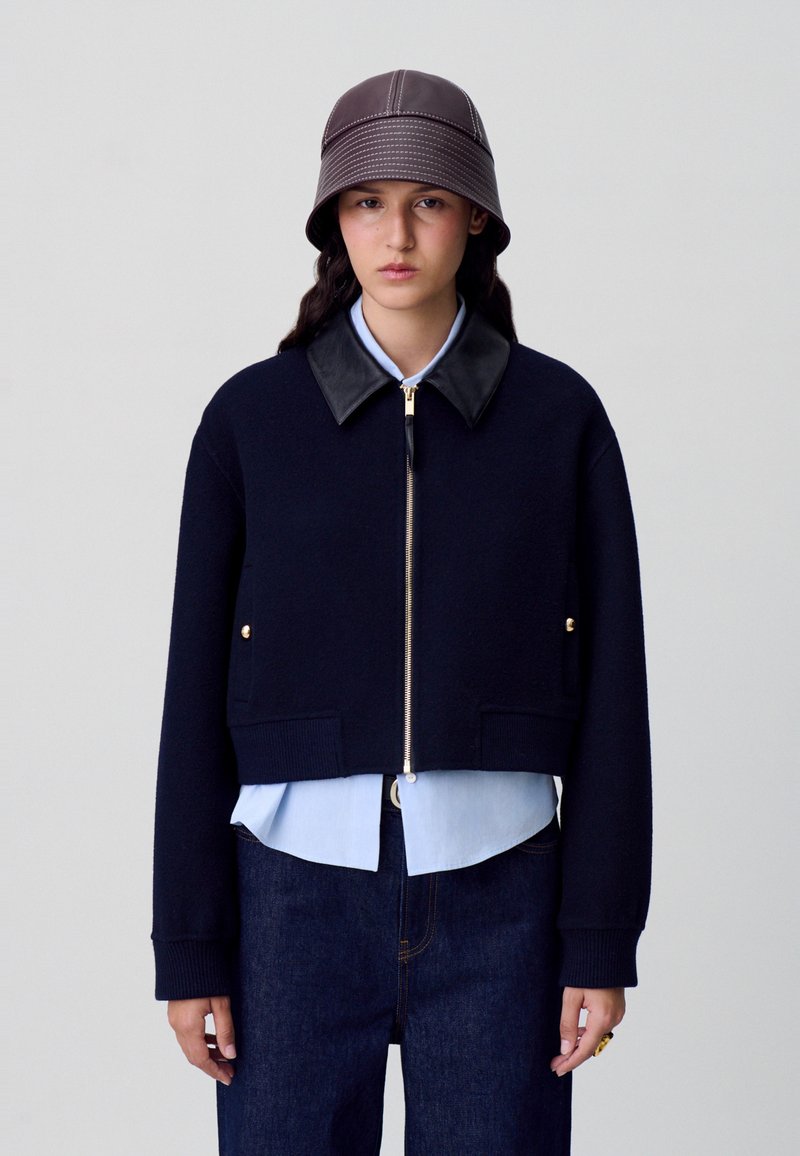 Chaqueta corta de color marino con cuello de piel, herrajes dorados y cierre de cremallera, superpuesta a una camisa azul clara y jeans de mezclilla oscura. Sombrero tipo bucket marrón.