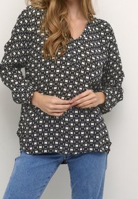 Kobieta w długim rękawie, nosząca czarno-białą bluzkę w geometryczny wzór, z niebieskimi dżinsami, z rękami złożonymi na talii, na jednolitym tle.