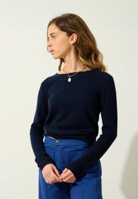 Maritimer gestrickter Pullover mit langen Ärmeln und weitem Ausschnitt, kombiniert mit blauen Hosen, die über vordere Taschen und ein markantes Knopfdetail verfügen.