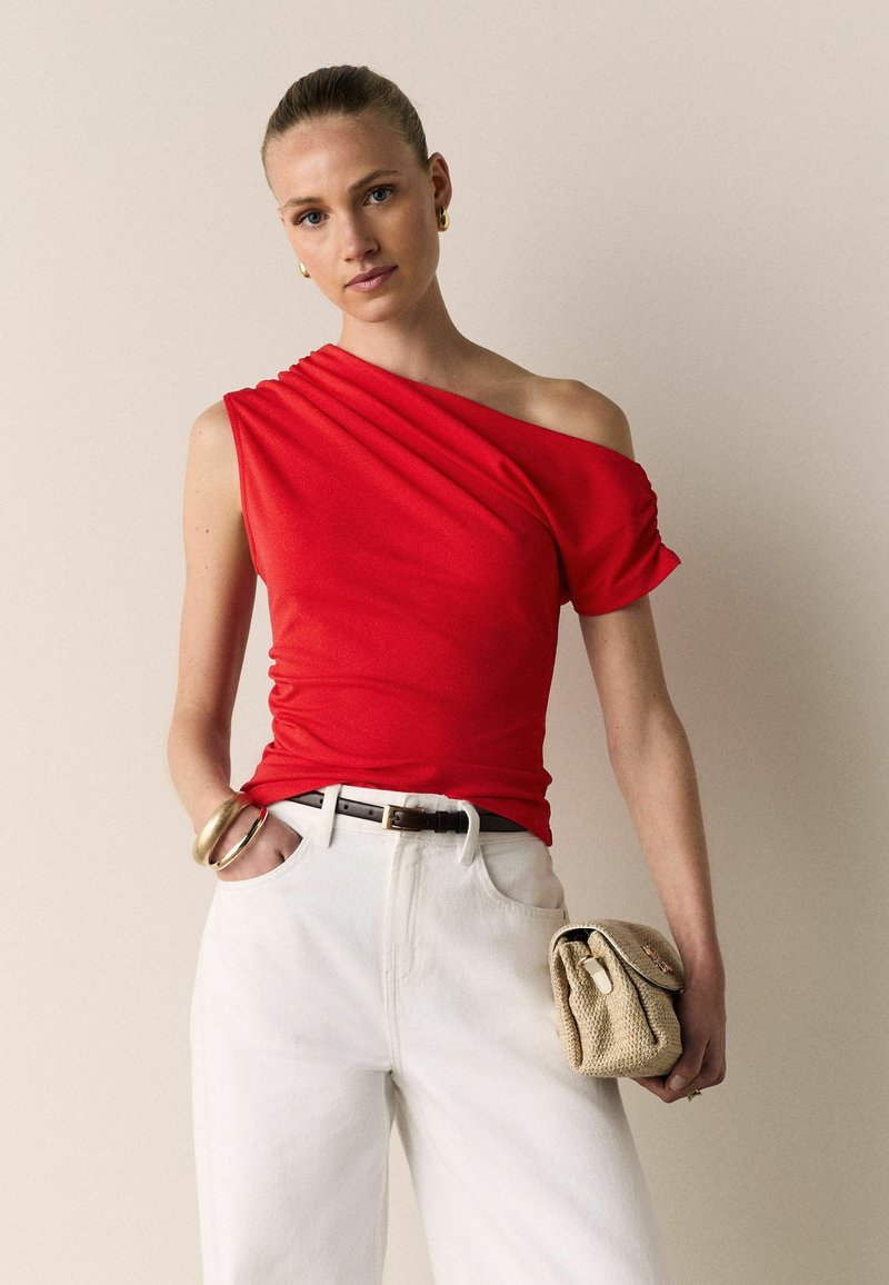 Femme portant un haut rouge asymétrique, un pantalon blanc taille haute avec une ceinture noire, des bracelets dorés, des boucles d'oreilles et tenant une pochette beige tressée.