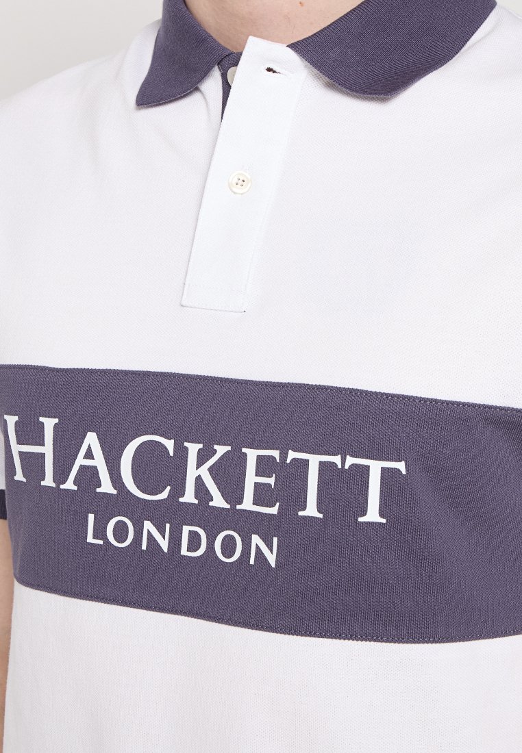 Polo à manches courtes présentant un corps blanc et une bande horizontale bleu marine, affichant le texte "HACKETT LONDON" en blanc au-dessus de la bande.