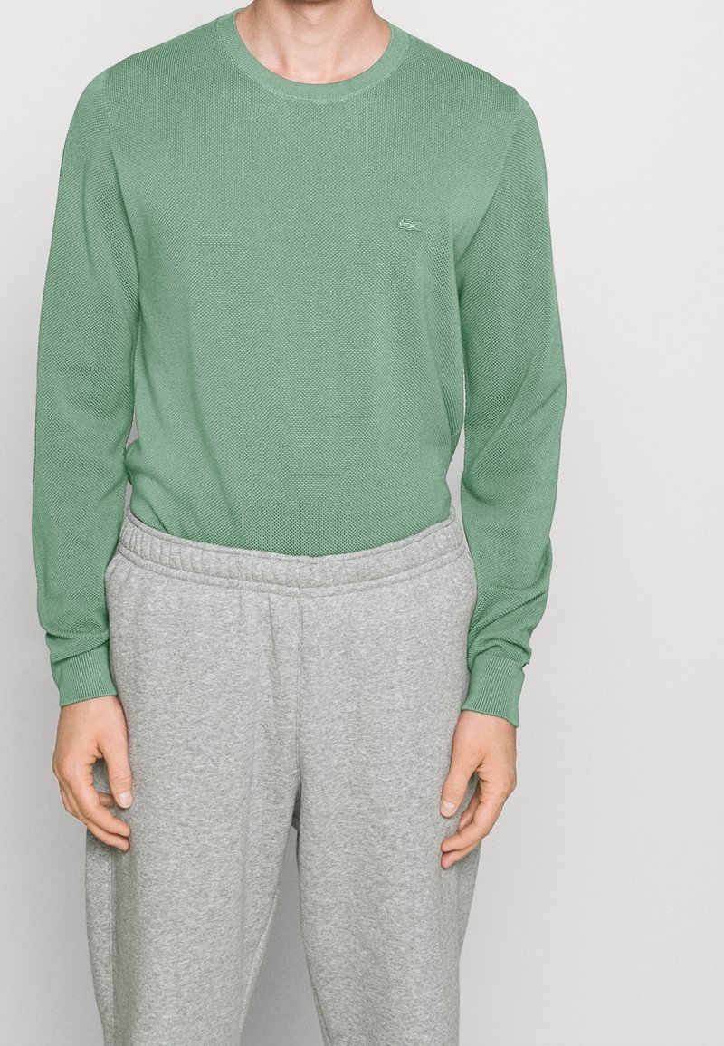 Zielona, długorękawowa dzianinowa koszulka z okrągłym dekoltem i fakturowanej tkaniny, zestawiona z szarymi dresami z elastycznym pasem.