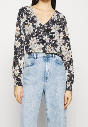 Vrouw in een blauw bloemen blouse met lange mouwen en V-hals, ingestopt in een hoge taille lichtblauwe jeans, staand tegen een effen achtergrond.