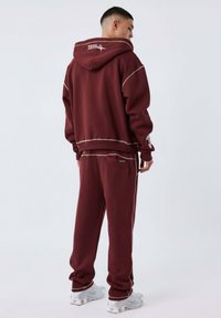 Set di felpa e pantaloni della tuta bordeaux, realizzato in materiale morbido, con cuciture a contrasto. Presenta un cappuccio e tasche laterali.