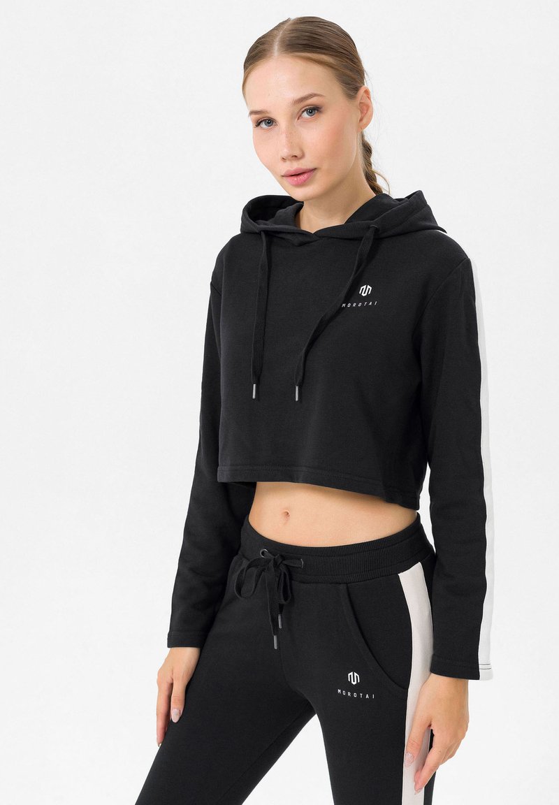 Felpa nera corta con cappuccio e lacci, caratterizzata da un logo sul petto. Abbinata a pantaloni sportivi neri con pannelli bianchi sui lati e tasche.