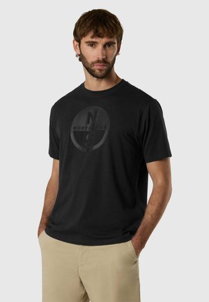 Giovane uomo con capelli castani e barba, indossa una t-shirt nera con il logo North Sails e pantaloni beige, mani in tasca, sfondo neutro.