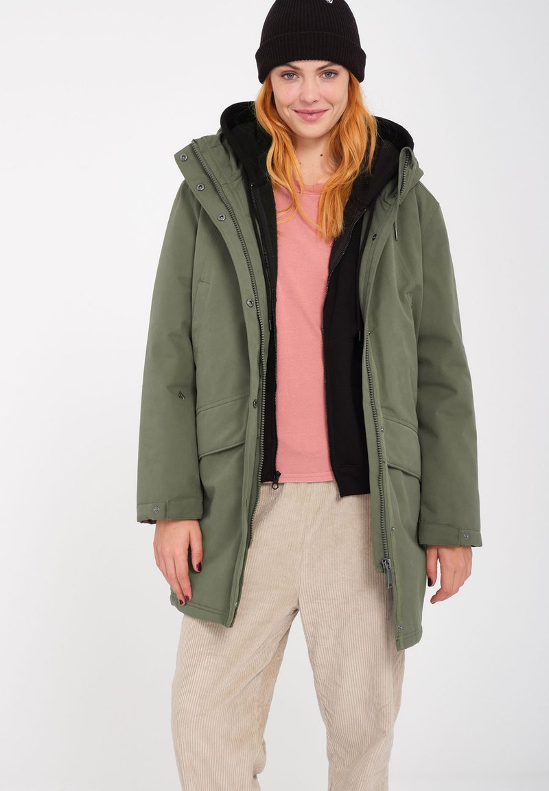 Veste extérieure verte avec une capuche et fermeture à boutons, superposée à une veste intérieure noire et un haut rose clair. Comprend deux poches avant.