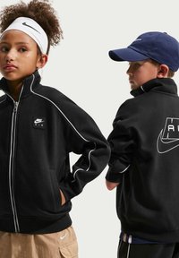 Δύο παιδιά φορούν μαύρα μπουφάν Nike Air· το κορίτσι κοιτάει προς τα εμπρός με άσπρη κορδέλα στο κεφάλι, ενώ το αγόρι κοιτάζει πλευρικά φορώντας σκούφο σκούρο μπλε.