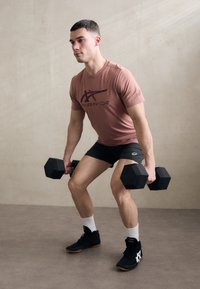 Roze t-shirt met korte mouwen en een donkere grafische print, zwarte shorts en zwarte sportschoenen. Hij houdt hexagonale dumbbells vast. Lichte neutrale achtergrond.