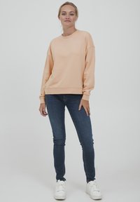 Ferskenfarget oversized sweatshirt med ribbestrikkede mansjetter, kombinert med blå skinny jeans og hvite joggesko. Glatt tekstur og uformell design.