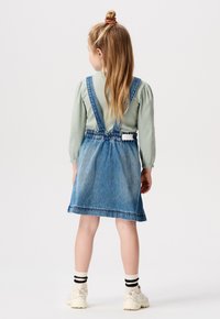 Denim-Overallkleid mit verstellbaren Trägern, elastischem Bund und gestuftem Rock. Langärmliges, geripptes grünes Oberteil, weiße Sneakers und gestreifte Socken.