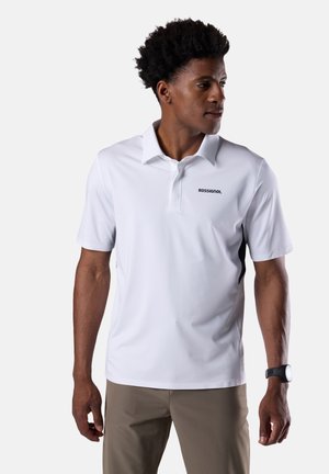 Homme aux cheveux bouclés foncés portant un polo blanc Rossignol, un pantalon kaki et une montre noire au poignet, regardant vers la droite.