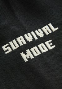 Tela negra con texto bordado en blanco que dice "MODO DE SUPERVIVENCIA" en una fuente gruesa y cuadrada. La textura es suave y uniforme.