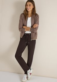 Jeune femme souriante, bras croisés, portant une veste zippée marron, un t-shirt blanc, un pantalon noir à cordon de serrage, et des baskets blanches.