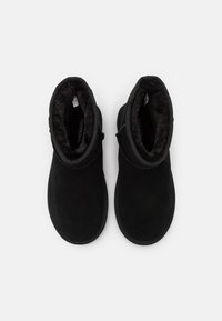 Botas de ante negras hasta el tobillo con un suave forro de pelo sintético. El diseño presenta una puntera redonda y detalles cosidos a lo largo del borde superior.