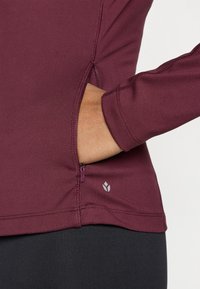 Tumši sarkana garroku krekls ar sānu rāvējslēdzēja kabatu, gludu audumu, pieguļošu dizainu un kontrastējošu pelēku logotipu pie apakšmala.