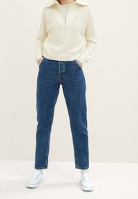 Pullover a maglia testurizzata color crema, con colletto ampio e chiusura a zip, abbinato a jeans blu a vita alta e sneakers bianche.