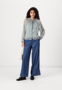 Helles blaues Bluse mit floralem Muster, gerafftem Rundhalsausschnitt, Knopfverschluss und transparenten langen Ärmeln; kombiniert mit weit geschnittenen Denim-Hosen und schwarzen Schuhen.