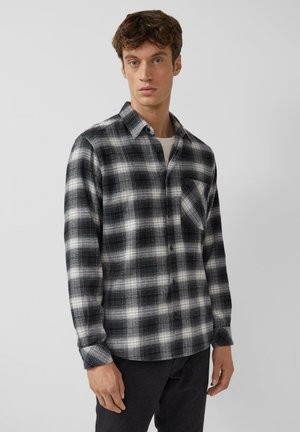 Młody mężczyzna w czarno-białej koszuli w kratę z długim rękawem na białym T-shircie, stojący na gładkim jasnym tle.