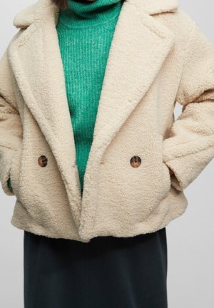 Gros plan d'une personne portant un manteau teddy crème avec des boutons marron sur un pull col roulé côtelé vert et une jupe noire. Mains dans les poches.
