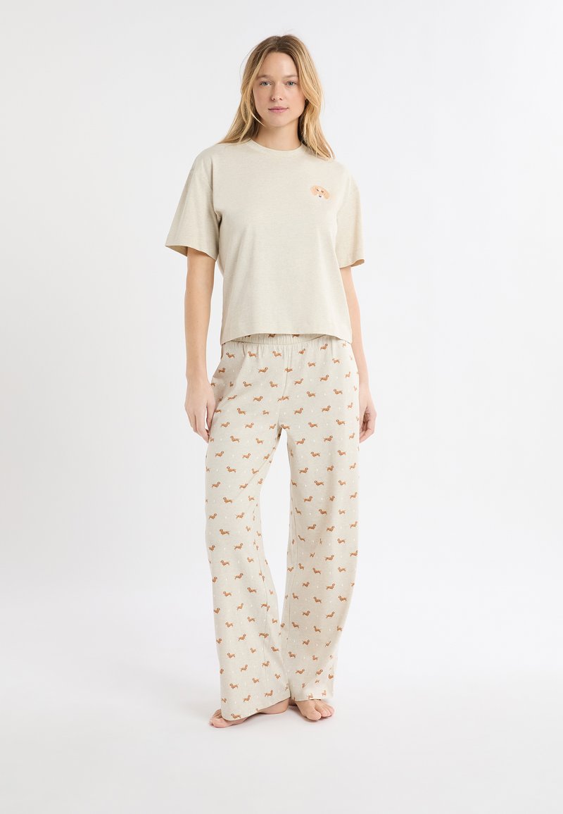 Beiges Kurzarmhemd mit einem verspielten Design auf der Brust, kombiniert mit hellen Hosen mit einem wiederholten Tiermuster.