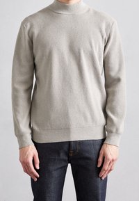 Pull gris clair en maille côtelée avec col montant et manches longues. Porté avec un jean foncé, présentant une coupe décontractée et un tissu texturé.