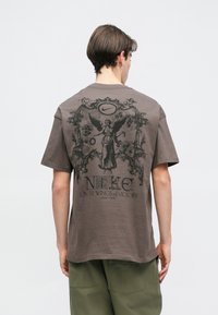 Καφέ βαμβακερό μπλουζάκι με μεγάλο μαύρο γραφικό σχέδιο στην πλάτη, που απεικονίζει έναν άγγελο με περίπλοκα στοιχεία και το λογότυπο της Nike από πάνω.