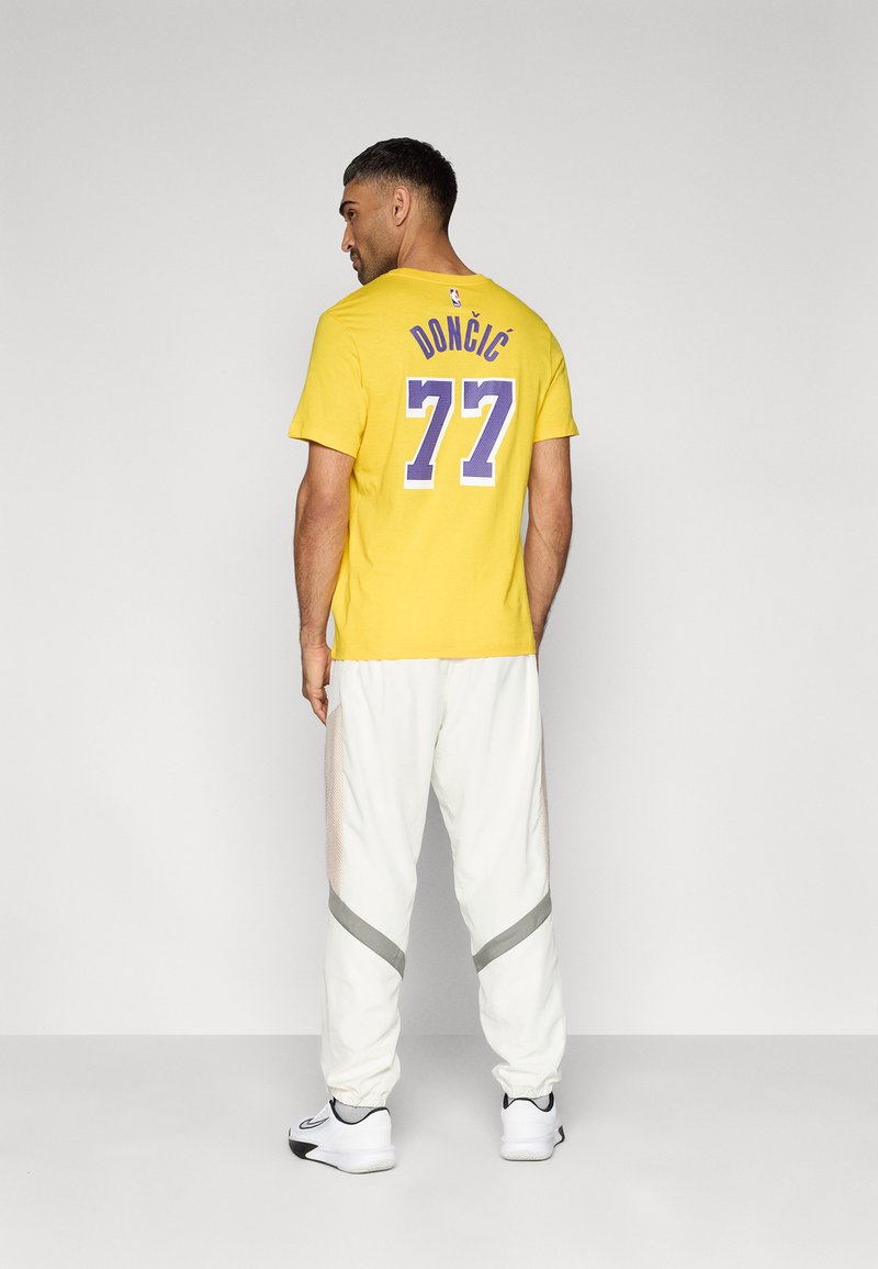 Gelbes Baumwoll-T-Shirt mit "DONČIĆ" und der Nummer "77" in Lila. Weiße Hose mit Netz-Einsätzen und grauen Akzenten. Sneakers mit schwarzen und weißen Details.