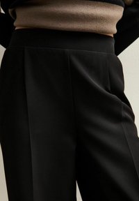 Pantalones negros ajustados con una textura suave, pliegues en la parte delantera y una cinturilla ancha, combinados con un suéter marrón por encima.