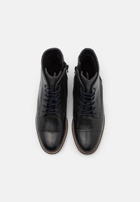 Bottes cuir noir à lacets montant à la cheville, avec fermeture éclair latérale, texture lisse, bout renforcé et surpiqûres marron contrastantes sur la semelle.