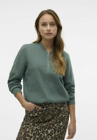 Veste à fermeture éclair verte avec un col rond, des poignets côtelés et des poches latérales, associée à une jupe à imprimé léopard marron.