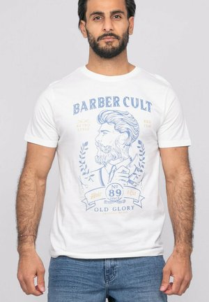 Homme barbu portant un t-shirt blanc avec une illustration vintage de barbier et le texte "Barber Cult", debout devant un fond uni.