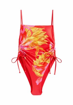 Žena v červeno-žltých kvetovaných jednodielnych plavkách upravuje bočné zaväzovanie vonku, v pozadí je zelené listnaté pozadie.